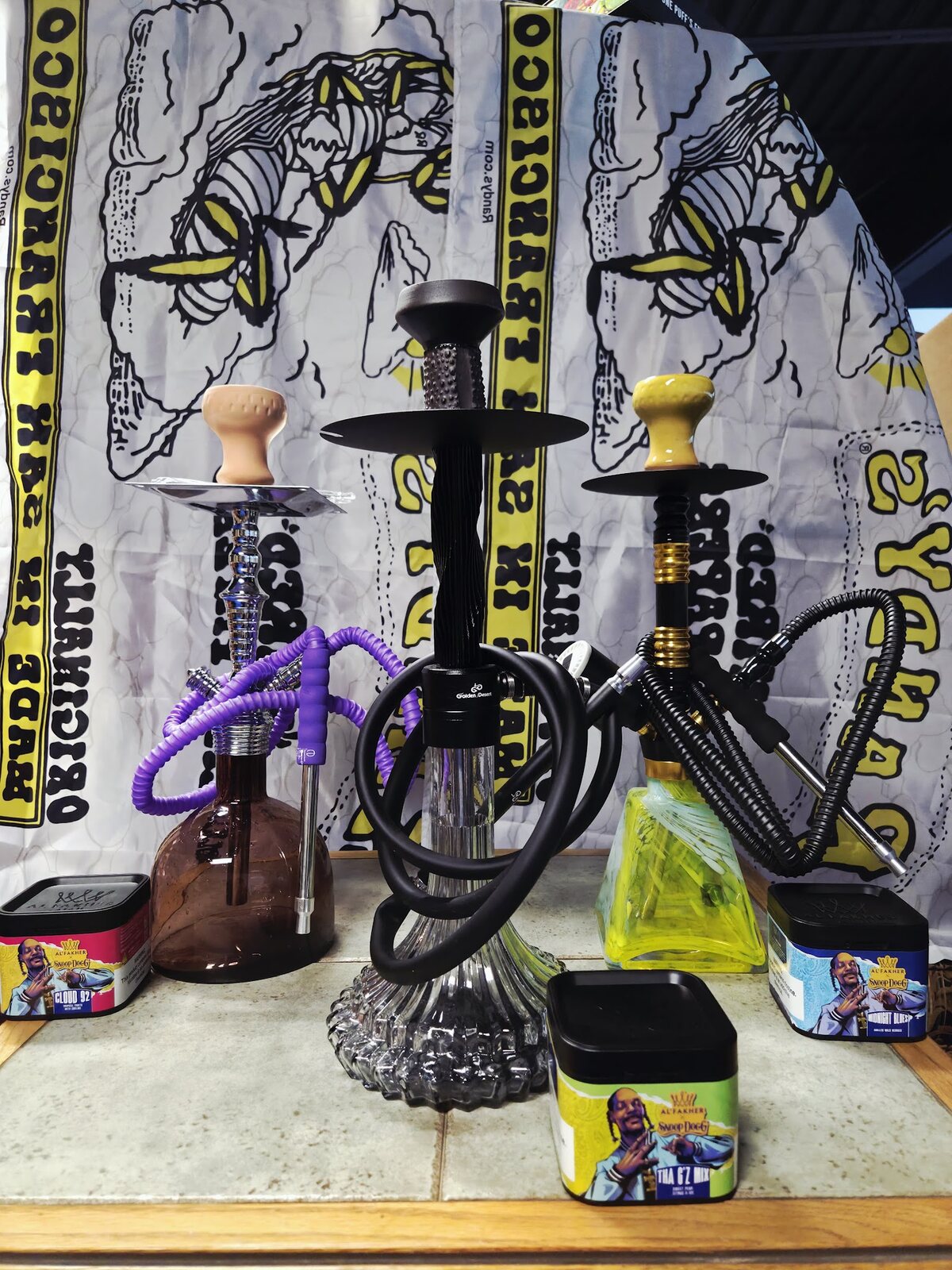 Three hookahs on the counter at Rafi Smoke Shop with Al Fakher Snoop Dogg shisha tins in front of a Randy's rolling papers banner.