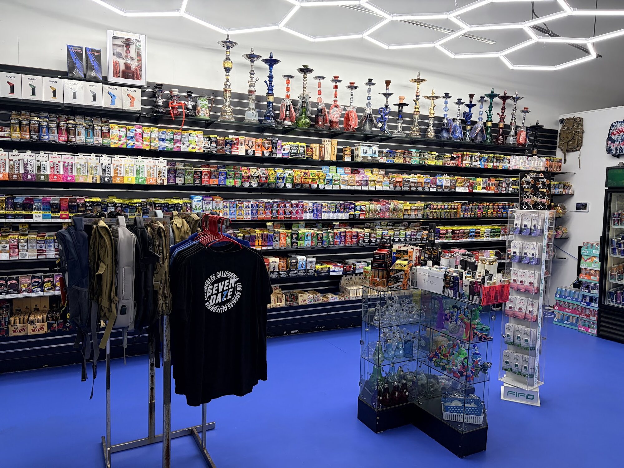 Inside Rafi Smoke Shop: hookahs along the top rail, wraps and rolling papers down the side wall, a glass case of hand pipes in the middle of the blue floor, under hexagonal LED ceiling lights.