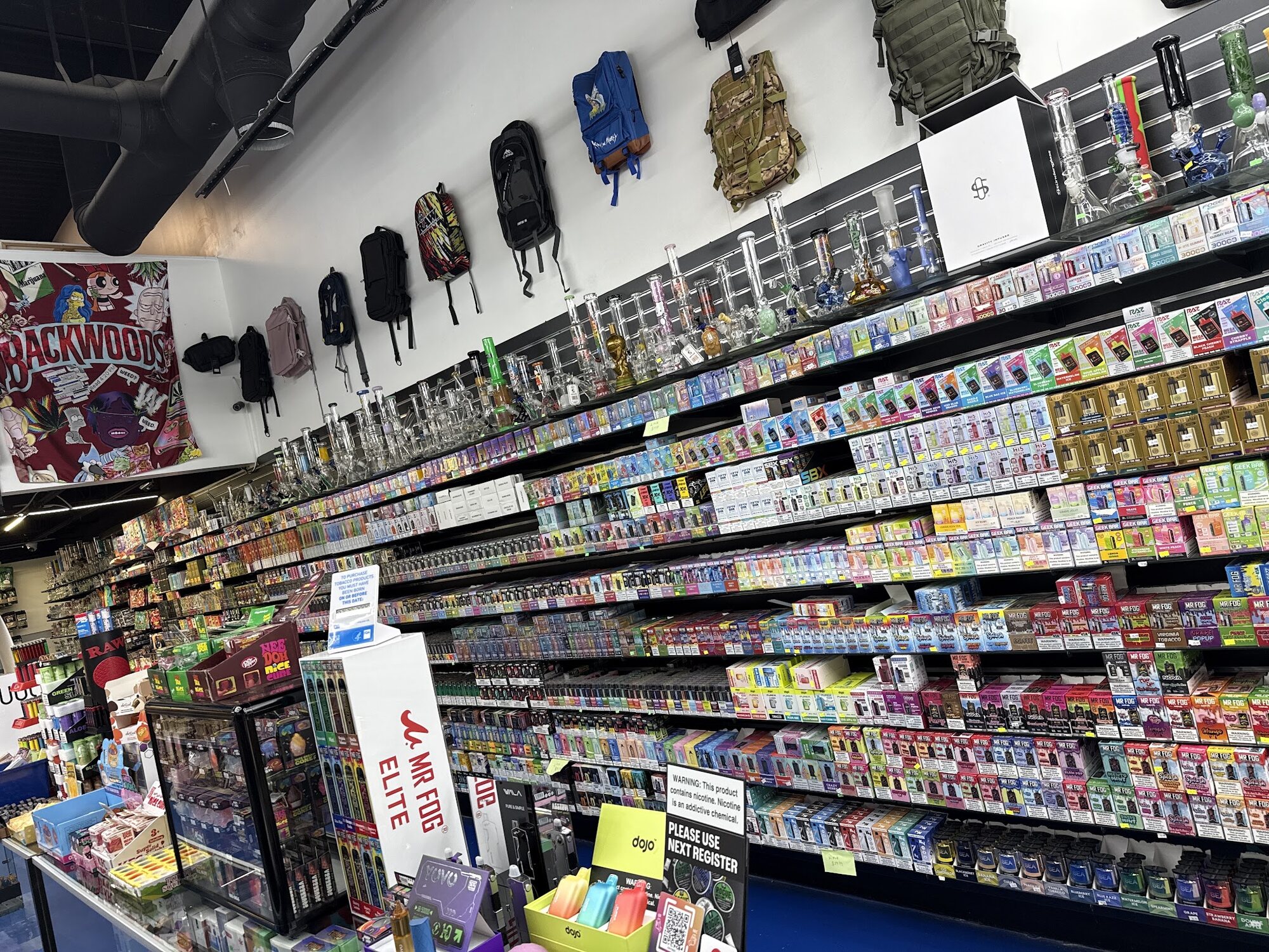 The back wall at Rafi Smoke Shop, stacked four rows high with disposable vapes, with water pipes lined along the shelf above.